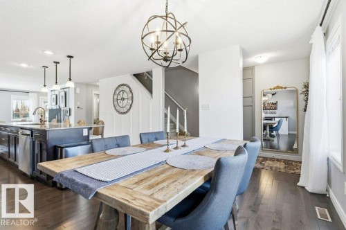 This open-concept dining area features hardwood floors, a large rustic dining table, and a contemporary chandelier - 16720 15 Avenue, Edmonton, AB - Indoor Photo Showing Dining Room