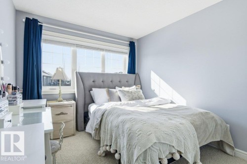 This room features a large window with blue curtains, light blue walls, and light-colored carpeting - 16720 15 Avenue, Edmonton, AB - Indoor Photo Showing Bedroom