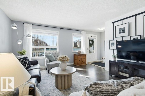 The living area features light-colored walls, dark hardwood flooring, and a window with white curtains - 16720 15 Avenue, Edmonton, AB - Indoor Photo Showing Living Room