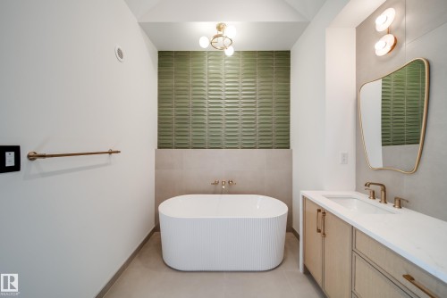 This bathroom features a freestanding tub, a vanity with a white countertop and gold-toned fixtures, a decorative mirror, and an accent wall with textured green tiles - 13804 85 Avenue, Edmonton, AB - Indoor Photo Showing Bathroom