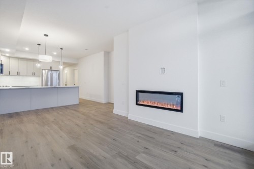 Open concept living area featuring light wood-style flooring, a modern electric fireplace, and a kitchen with light-colored cabinetry, a stainless steel refrigerator, and pendant lighting - 516 260 Bellerose Dr, St. Albert, AB - Indoor With Fireplace