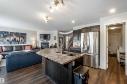Open concept kitchen and living area featuring dark wood flooring, a central kitchen island with an undermount sink, dark kitchen cabinetry, and a stainless steel refrigerator - 