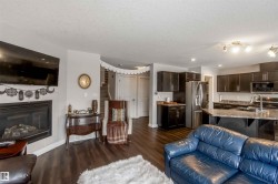 This open-concept living space features dark wood-style flooring, a fireplace with a light-colored surround, and a kitchen with dark cabinetry, stainless steel appliances, and a central island with a sink - 