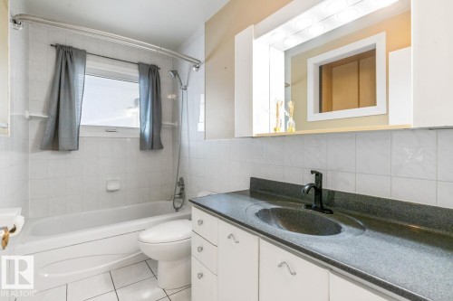 Bathroom featuring a white bathtub with a window, white tiled walls, and a vanity with a dark countertop and white cabinetry - 7324 77 Street, Edmonton, AB - Indoor Photo Showing Bathroom