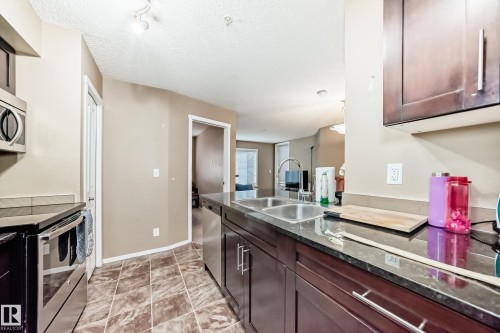 The kitchen features dark wood cabinetry, a double basin stainless steel sink, and dark countertops - 116 5810 Mullen Place, Edmonton, AB - Indoor Photo Showing Kitchen With Double Sink