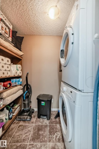 Laundry area featuring a stacked washer and dryer, tiled flooring, and a built-in shelving unit - 116 5810 Mullen Place, Edmonton, AB - Indoor Photo Showing Laundry Room