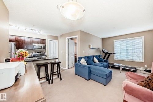This open-concept living area features light-colored carpeting, neutral painted walls, and a large window with blinds - 116 5810 Mullen Place, Edmonton, AB - Indoor Photo Showing Living Room