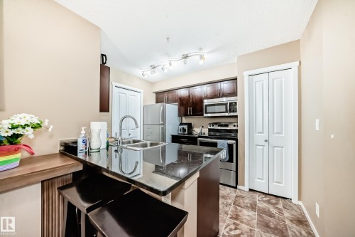 The kitchen features dark wood cabinetry, stainless steel appliances, and a granite countertop island with seating - 116 5810 Mullen Place, Edmonton, AB - Indoor Photo Showing Kitchen With Double Sink With Upgraded Kitchen