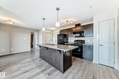 The kitchen features dark cabinetry, black appliances, and a central island with a sink and stone-look countertop - 332 344 Windermere Road, Edmonton, AB - Indoor Photo Showing Kitchen With Upgraded Kitchen