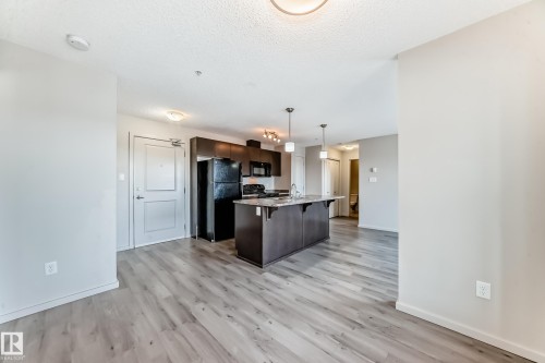 The property features light-toned flooring throughout, a kitchen with dark cabinetry and black appliances, a central kitchen island with a sink, and pendant lighting - 332 344 Windermere Road, Edmonton, AB - Indoor Photo Showing Kitchen