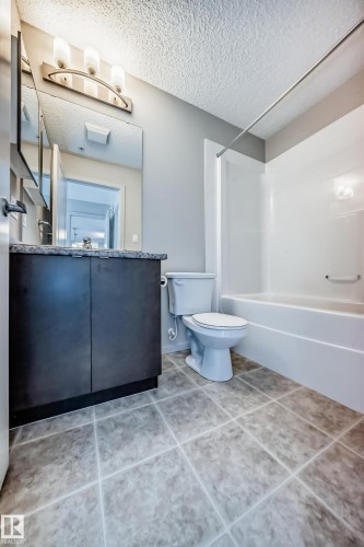 Bathroom featuring a vanity with dark cabinetry and a light-colored countertop, a toilet, and a white bathtub with a shower rod - 332 344 Windermere Road, Edmonton, AB - Indoor Photo Showing Bathroom