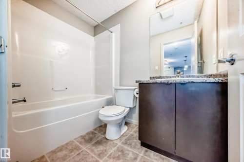 Bathroom featuring a built-in bathtub with shower, a vanity with a dark wood cabinet and stone countertop, and tiled flooring - 332 344 Windermere Road, Edmonton, AB - Indoor Photo Showing Bathroom