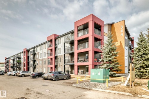 Exterior view of a multi-story building featuring balconies with railings, modern architectural design elements in red and orange, and an adjacent paved parking area - 332 344 Windermere Road, Edmonton, AB - Outdoor With Balcony With Facade