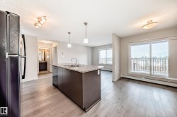 This kitchen and living area features a kitchen island with a double sink and pendant lighting, light hardwood floors, and a balcony with a metal railing - 