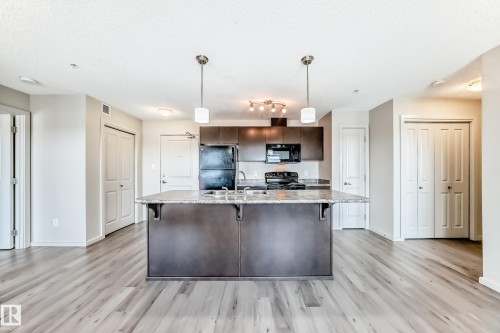 Kitchen and living area featuring a large island with a light-colored countertop, dark wood cabinetry, and stainless steel appliances - 332 344 Windermere Road, Edmonton, AB - Indoor Photo Showing Kitchen