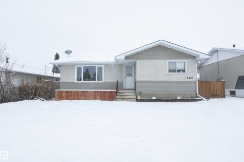 This property features a single-story home with light-colored stucco and horizontal siding, a light-colored entry door, and a front yard with a wooden fence - 4420 56 Avenue, Barrhead, AB - Outdoor