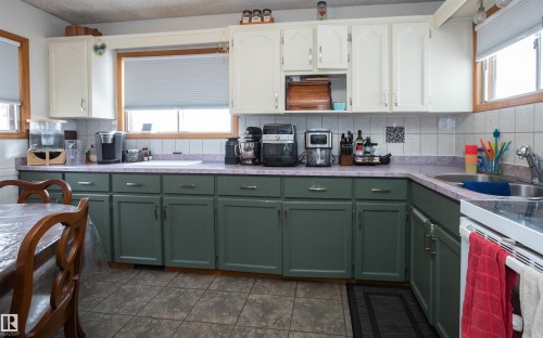 The kitchen features white upper cabinetry and green lower cabinetry, a light-colored tile backsplash, and a double basin stainless steel sink - 4420 56 Avenue, Barrhead, AB - Indoor Photo Showing Kitchen With Double Sink