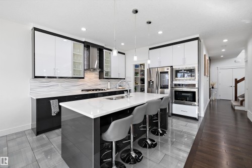 Modern kitchen featuring a large island with a white countertop and seating for three, sleek two-tone cabinetry, stainless steel appliances, and a unique textured backsplash - 6303 169 Avenue, Edmonton, AB - Indoor Photo Showing Kitchen With Stainless Steel Kitchen With Double Sink With Upgraded Kitchen