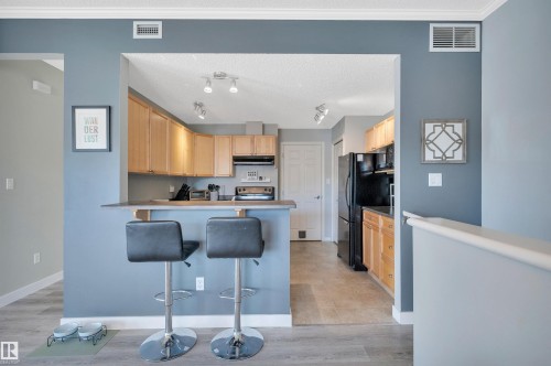 The property features a kitchen with light wood cabinetry, black appliances, and a breakfast bar with two bar stools - 106 3040 Spence Wynd, Edmonton, AB - Indoor Photo Showing Kitchen