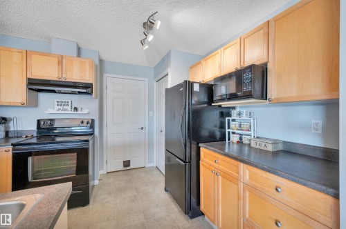 The kitchen features light wood cabinetry, black appliances including a refrigerator, microwave, and range, and dark countertops - 106 3040 Spence Wynd, Edmonton, AB - Indoor Photo Showing Kitchen