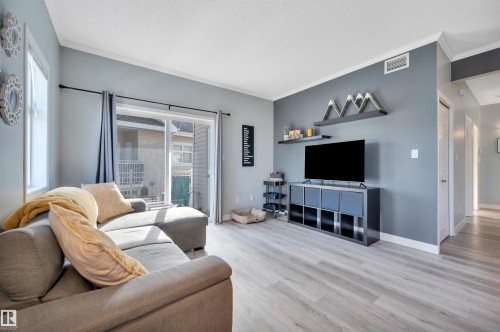 This living area features light-toned flooring, a gray accent wall, and a sliding glass door leading to a balcony - 106 3040 Spence Wynd, Edmonton, AB - Indoor Photo Showing Living Room