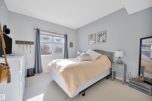 This room features a window with curtains, a bed with a fabric headboard, and light-colored carpeting - 106 3040 Spence Wynd, Edmonton, AB - Indoor Photo Showing Bedroom