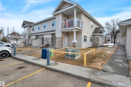 The property features a two-story exterior with light-colored siding and stone accents, a covered balcony with white railings, and a paved parking area - 106 3040 Spence Wynd, Edmonton, AB - Outdoor With Balcony