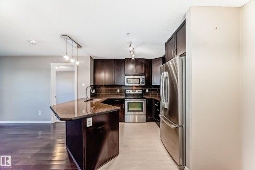 The kitchen features dark wood cabinetry, a granite countertop island with a sink, and stainless steel appliances - 208 2204 44 Ave, Edmonton, AB - Indoor Photo Showing Kitchen With Upgraded Kitchen