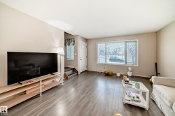 Spacious living area featuring light-toned walls, wood-look flooring, and a large window providing views of the outdoors - 