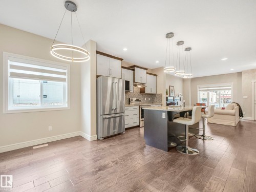 This open-concept living space features light wood-look flooring, a kitchen island with seating, pendant lighting, and stainless steel appliances - 2 10319 120 Street, Edmonton, AB - Indoor