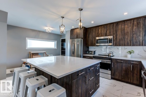 The kitchen features a spacious island with a white countertop, dark wood cabinetry, stainless steel appliances, and a white marble-patterned backsplash - 9526 109A Avenue, Edmonton, AB - Indoor Photo Showing Kitchen