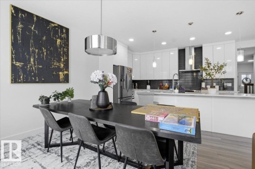 Open-concept kitchen and dining area featuring a dark dining table with seating, a large contemporary pendant light, and a kitchen with white cabinetry and stainless steel appliances - 16624 32 Avenue, Edmonton, AB - Indoor Photo Showing Dining Room