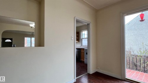 This area features dark wood flooring, a doorway leading to a kitchen with light wood cabinetry and a window, and a glass door opening to an outdoor patio with terracotta-style tiles and metal railings - 10839 109 Street, Edmonton, AB - Indoor Photo Showing Other Room