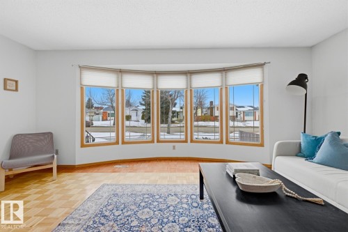 Living area featuring a prominent bay window with light-filtering blinds, light-toned parquet flooring, and a large area rug - 16108 100 Street, Edmonton, AB - Indoor Photo Showing Living Room