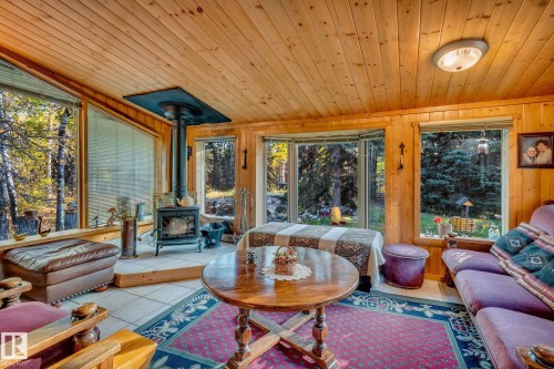 This inviting living area features extensive wood paneling on the walls and ceiling, a wood-burning stove, and large windows that provide views of the surrounding trees and yard - 60017 Range Road 42, Rural Barrhead County, AB - Indoor Photo Showing Living Room With Fireplace