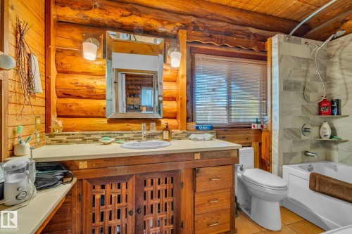 This bathroom features natural wood walls and ceiling, a vanity with a light-colored countertop and integrated sink, a mirror with wall-mounted lighting, a toilet, and a bathtub with a tiled shower surround - 60017 Range Road 42, Rural Barrhead County, AB - Indoor Photo Showing Bathroom