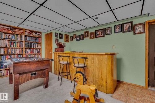 The property features a built-in wooden bar with two bar stools, a full wall of bookshelves, and a ceiling with a grid pattern - 60017 Range Road 42, Rural Barrhead County, AB - Indoor Photo Showing Other Room