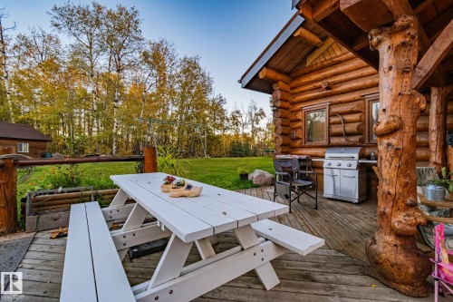 The property features an extensive wooden deck with a white picnic table, offering a view of a spacious lawn and mature trees - 60017 Range Road 42, Rural Barrhead County, AB - Outdoor With Deck Patio Veranda
