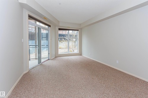 The room features light beige carpeted flooring, large windows, and a glass sliding door leading to a balcony with glass railings - 415 10503 98 Avenue, Edmonton, AB - Indoor Photo Showing Other Room