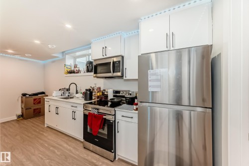 The kitchen features white cabinetry, stainless steel appliances including a refrigerator, oven, and microwave, a sink with a black faucet, and light-colored flooring - 12435 47 Street Nw, Edmonton, AB - Indoor Photo Showing Kitchen