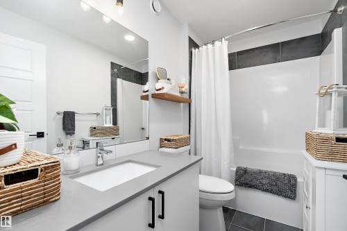 The bathroom features a vanity with a light gray countertop and an integrated sink, a large mirror, a shower-tub combination with a white shower curtain, and dark gray floor tiles - 20 Hemingway Crescent, Spruce Grove, AB - Indoor Photo Showing Bathroom