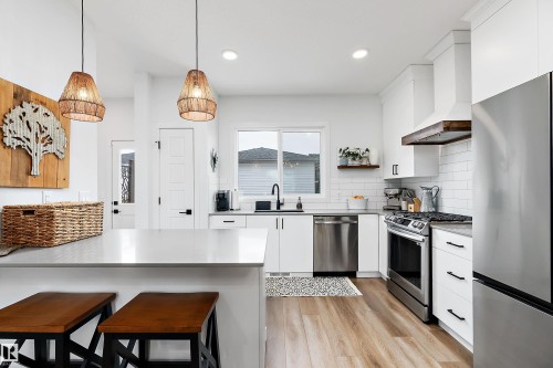 Modern kitchen featuring a kitchen island with seating, pendant lighting, stainless steel appliances, a white subway tile backsplash, and light wood flooring - 20 Hemingway Crescent, Spruce Grove, AB - Indoor Photo Showing Kitchen With Upgraded Kitchen