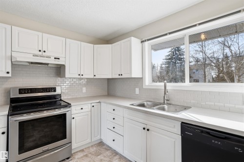 The kitchen features white cabinetry with black hardware, a stainless steel range, a double basin sink, and a light-colored subway tile backsplash - 18297 74 Avenue, Edmonton, AB - Indoor Photo Showing Kitchen With Double Sink