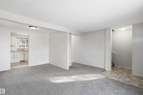 Spacious interior with light gray walls and gray carpet, featuring a kitchen with white cabinetry and a tiled backsplash, and a staircase with a white handrail - 18297 74 Avenue, Edmonton, AB - Indoor Photo Showing Other Room