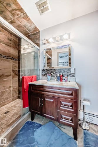 The bathroom features a shower with glass enclosure and tiled walls, a vanity with a light-colored countertop and dark wood cabinetry, and a mirrored wall fixture with sconce lighting - 12131 101 Street Nw, Edmonton, AB - Indoor Photo Showing Bathroom