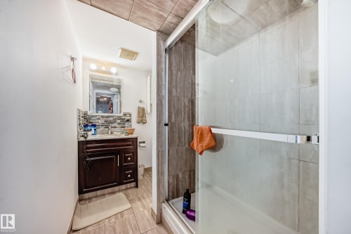 Bathroom featuring a dark wood vanity with a stone-tiled backsplash, a mirror with vanity lighting, and a glass-enclosed shower with light-toned wall tiles - 12131 101 Street Nw, Edmonton, AB - Indoor Photo Showing Bathroom