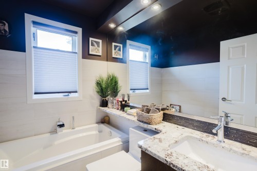 The bathroom features a built-in bathtub, dual windows with privacy blinds, and a vanity with a white granite countertop and an undermount sink - 10643 66 Avenue, Edmonton, AB - Indoor Photo Showing Bathroom