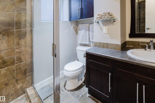 This bathroom features a shower with glass enclosure and stone-tiled walls, a dark wood vanity with a white sink and chrome faucet, and a toilet - 10643 66 Avenue, Edmonton, AB - Indoor Photo Showing Bathroom