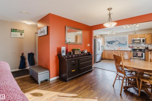 This interior view features hardwood flooring in the foreground, with a partial view of a kitchen that includes light wood cabinetry, stainless steel appliances, and a window above the sink - 8707 151 Avenue, Edmonton, AB - Indoor