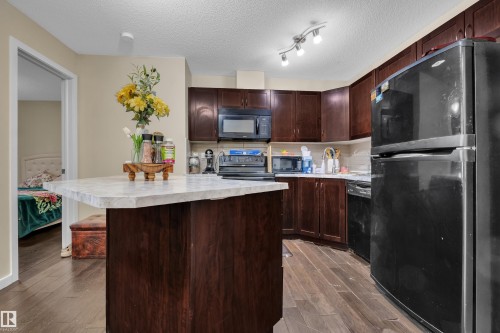 The kitchen features dark wood cabinetry, a central island with a light-colored countertop, and dark-finished appliances including a refrigerator, microwave, and dishwasher - 320 392 Silver Berry Road, Edmonton, AB - Indoor Photo Showing Kitchen